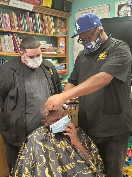 Two men wearing face masks are cutting a man 's hair in a barber shop