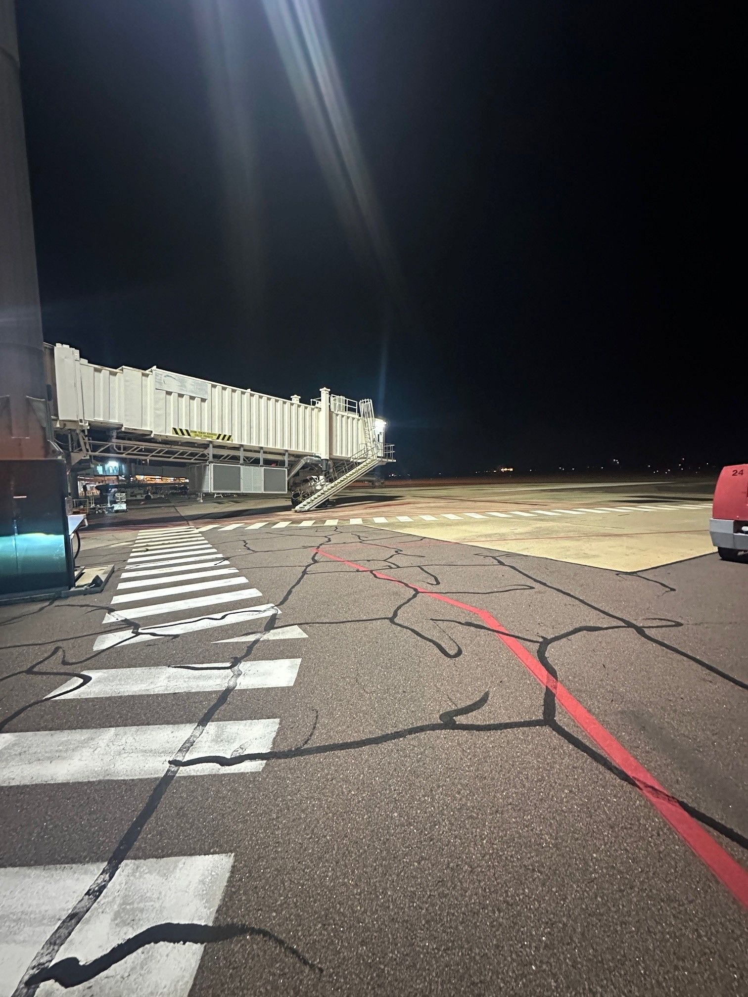 Empty nighttime runway with cracked pavement, painted markings, and parked vehicles beside a lit building