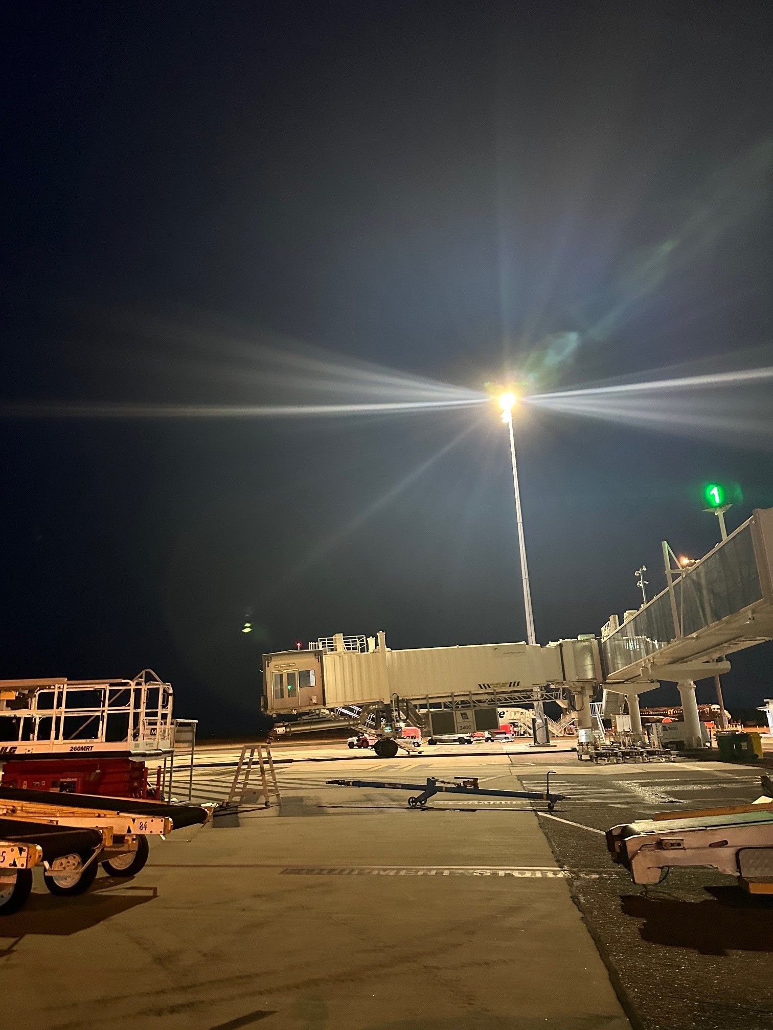 Nighttime airport tarmac with parked carts, cargo equipment, and a bright overhead light