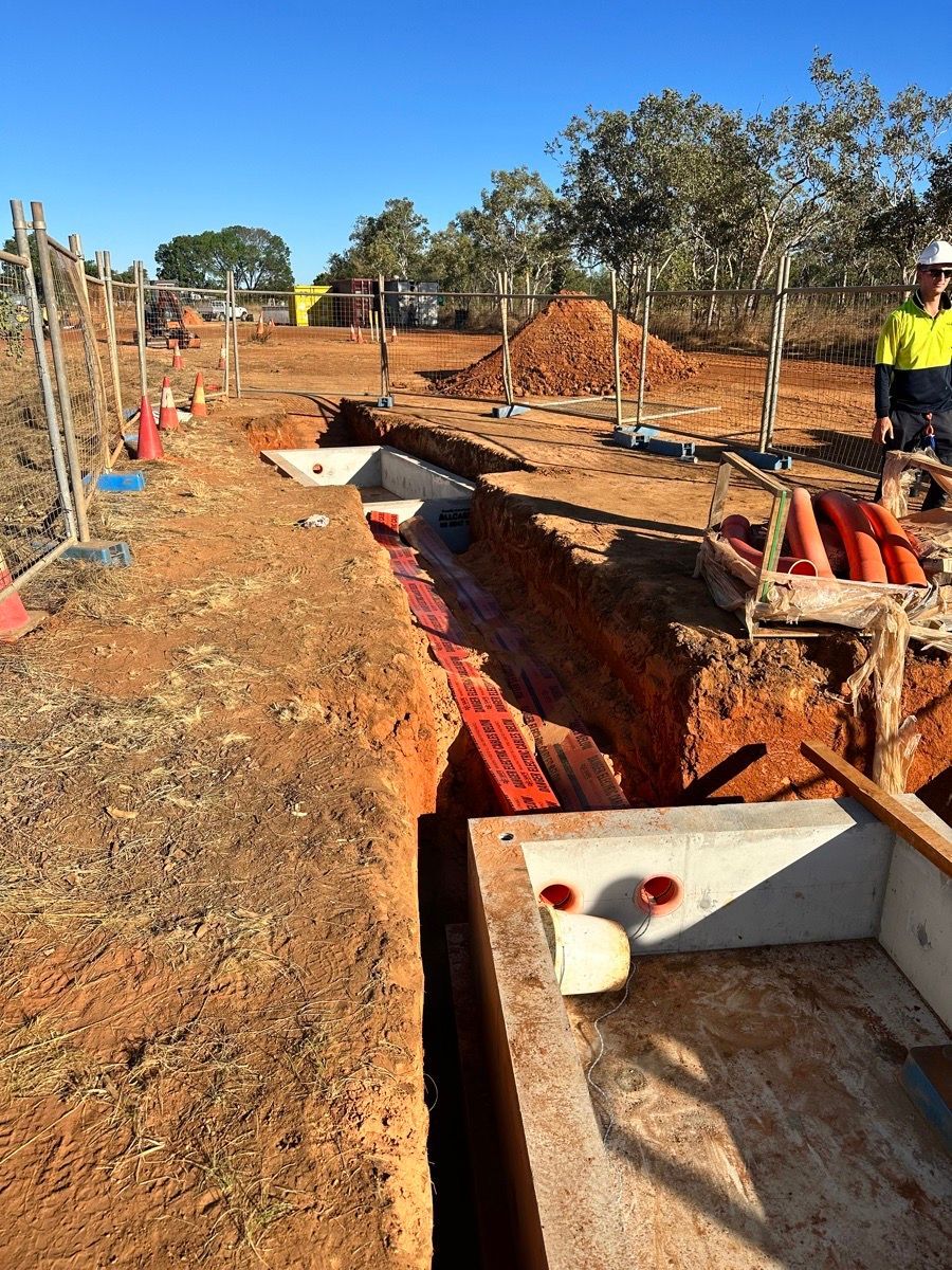 Construction site with open trench, red soil, and a worker in a high-visibility vest in the background