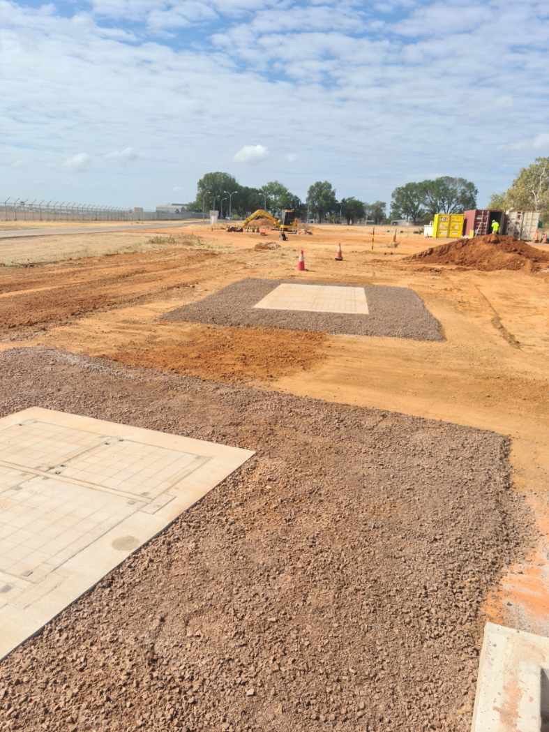 Construction site with dirt road, concrete slabs, and small workers in the distance under a cloudy sky