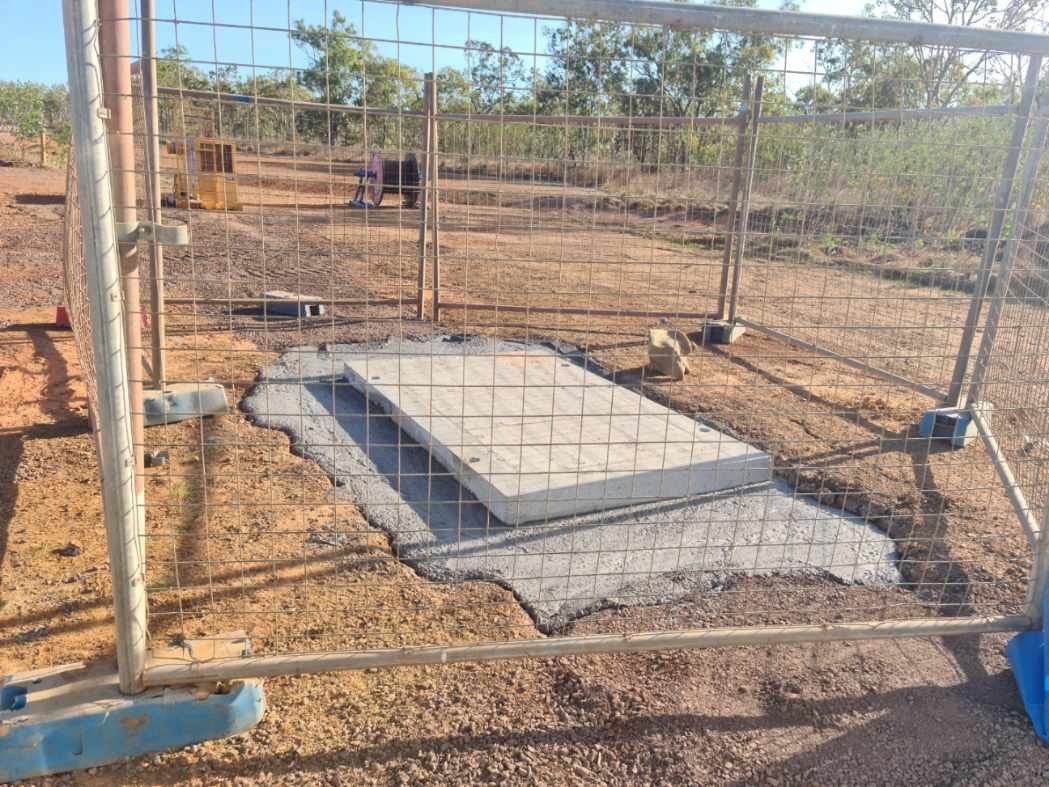 Concrete foundation slab under construction behind a wire fence in a dry, rural yard.