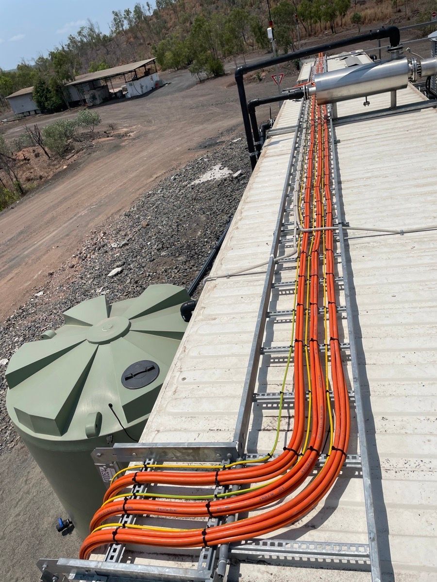 Orange cables run along a concrete path beside green tanks at an outdoor utility site.