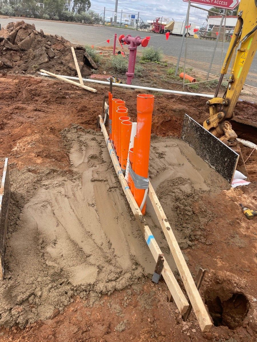 Construction trench with orange utility box, pipes, dirt piles, and an excavator arm nearby