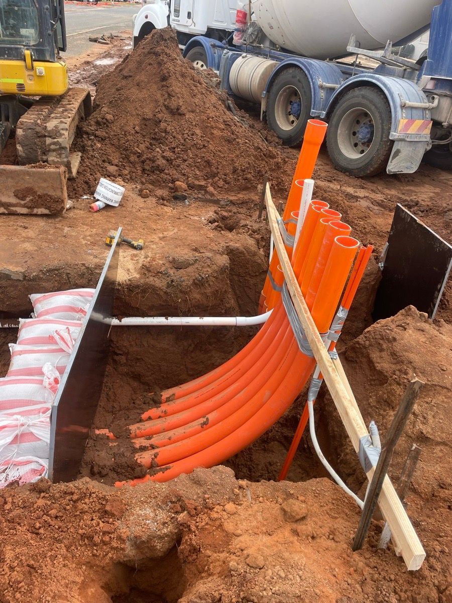 Construction trench with orange utility conduits being installed beside a concrete mixer truck and excavator.