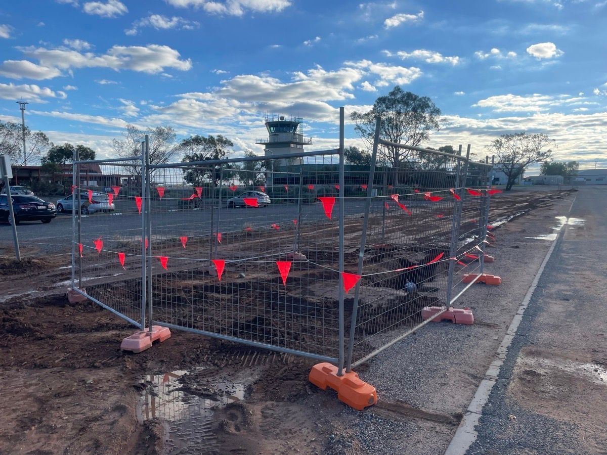 Fenced construction site with red flags on muddy ground under a cloudy sky.