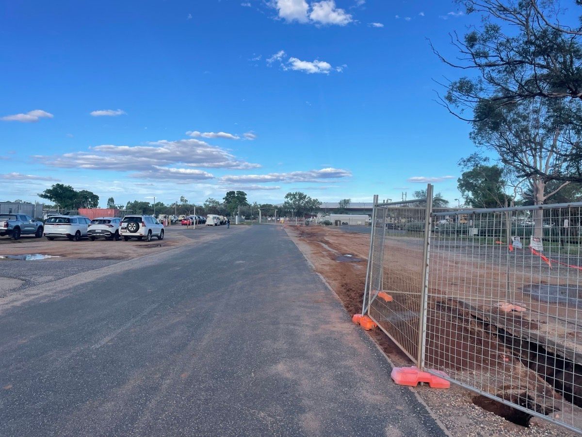 Dirt road beside chain-link fence and construction trench under a blue sky with parked vehicles on the left