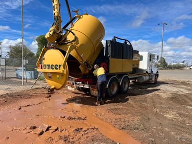 Yellow concrete mixer truck dumping wet concrete on a muddy construction site, worker standing beside it.