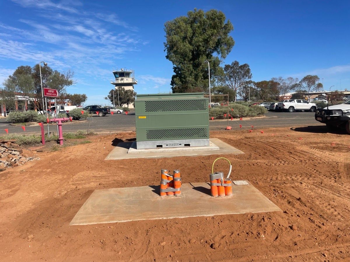 Construction site with utility boxes on concrete pads, orange cones, dirt ground, and vehicles in the background