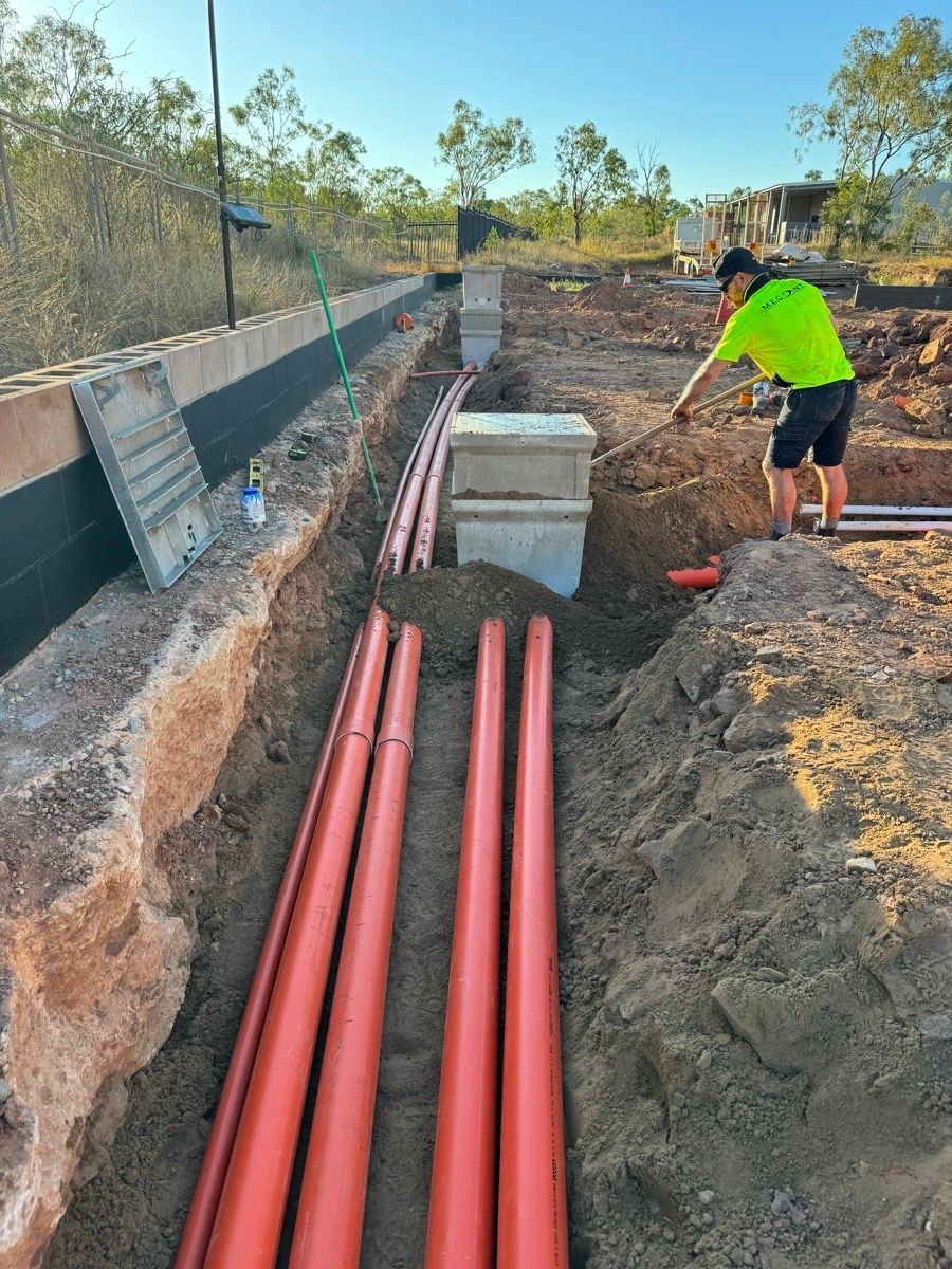 Worker at roadside trench installing red utility pipes beside a concrete wall