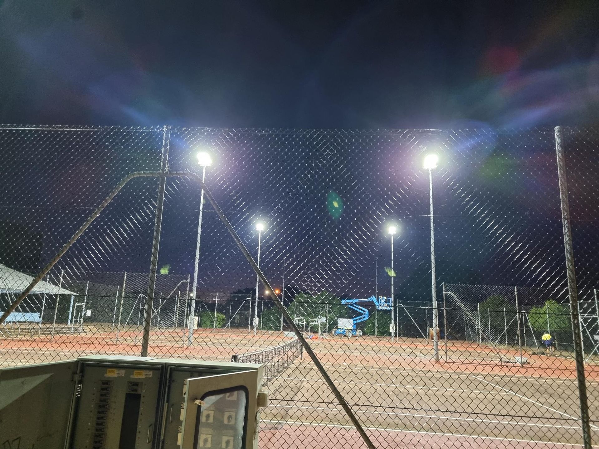 Floodlit tennis courts at night, viewed through a chain-link fence with a bench in the foreground