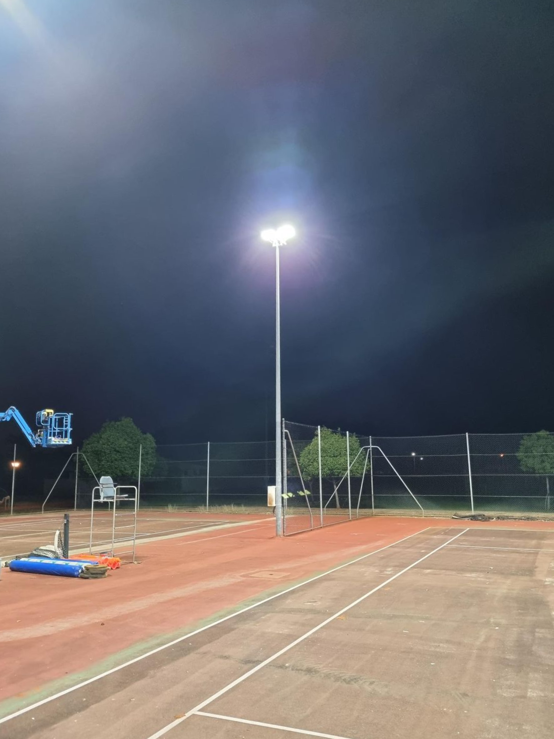 Floodlit outdoor tennis court at night with nets, fence, and blue equipment on the sideline