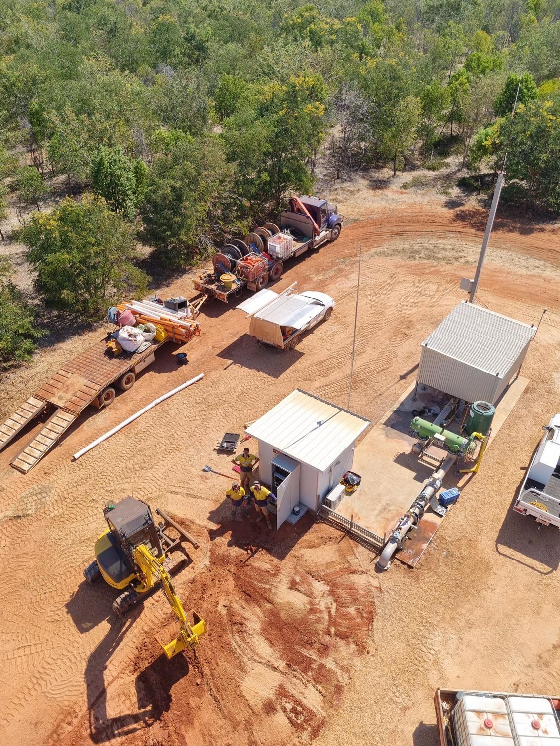 Aerial view of a construction site with heavy machinery, trailers, and cleared dirt beside dense trees