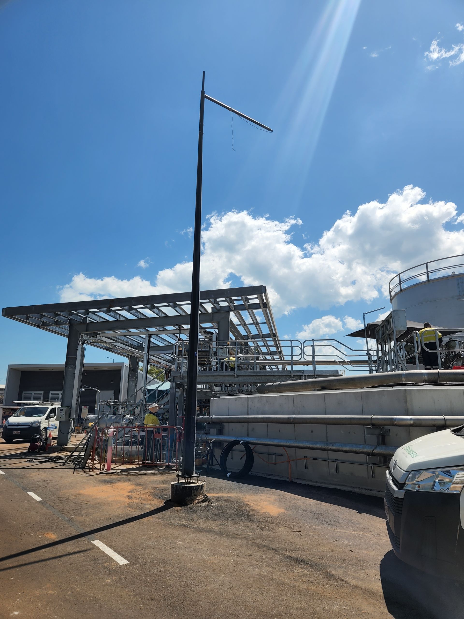 Industrial dock with metal structures, tanks, and a tall pole under a bright blue sky with clouds.