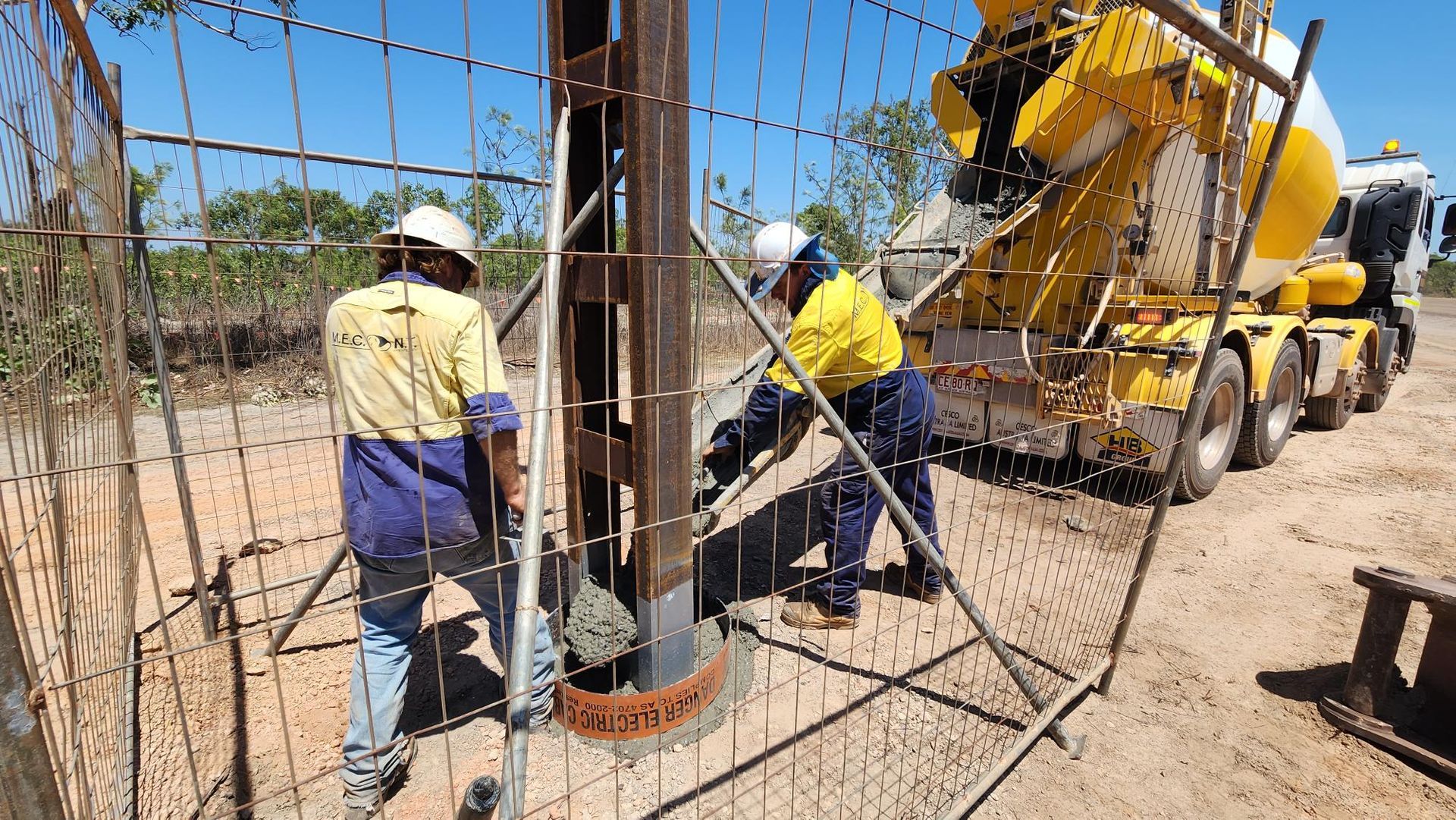 Workers operating a concrete mixer at a construction site behind a chain-link fence