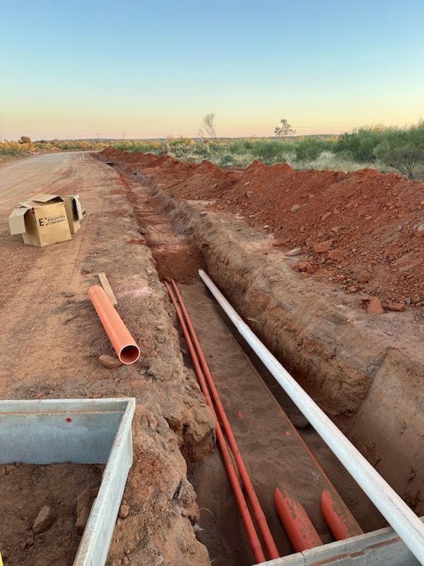Trench with orange and white utility pipes laid beside a dirt road at sunset.