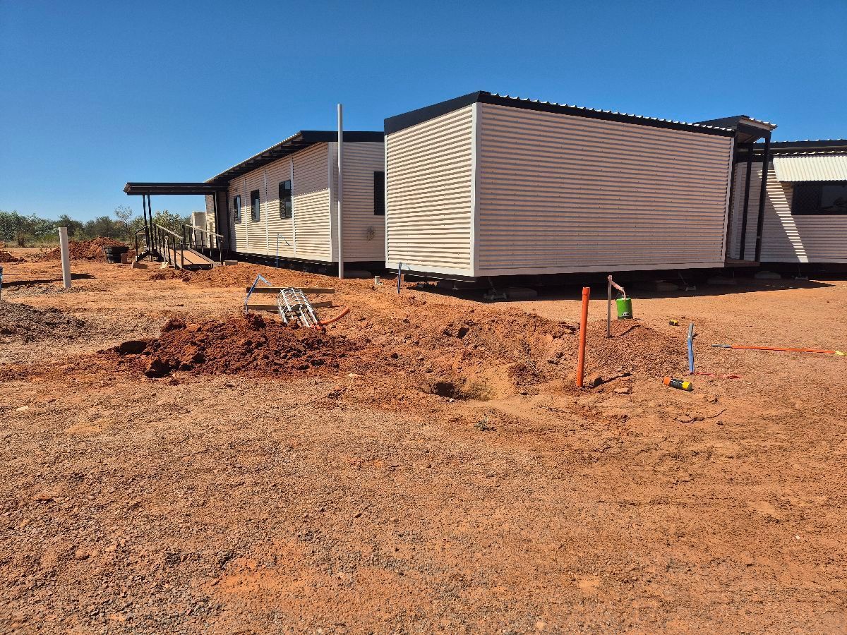 Two beige modular buildings on a dirt lot with utility stakes and a clear blue sky