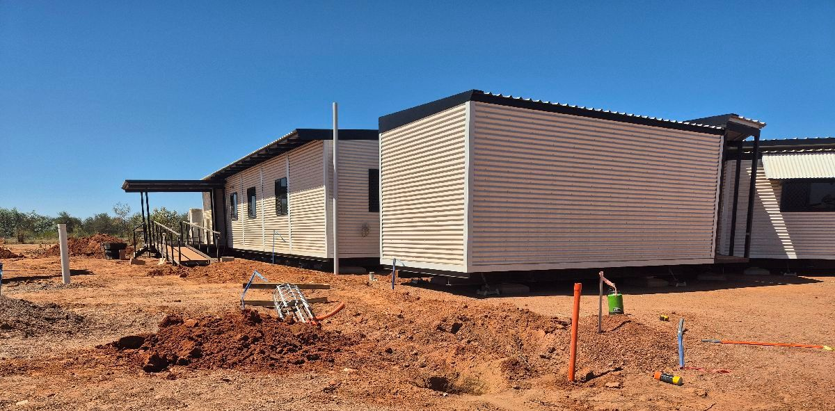 Two beige modular buildings on a dry dirt lot under a clear sky, with a utility pole and sparse vegetation.