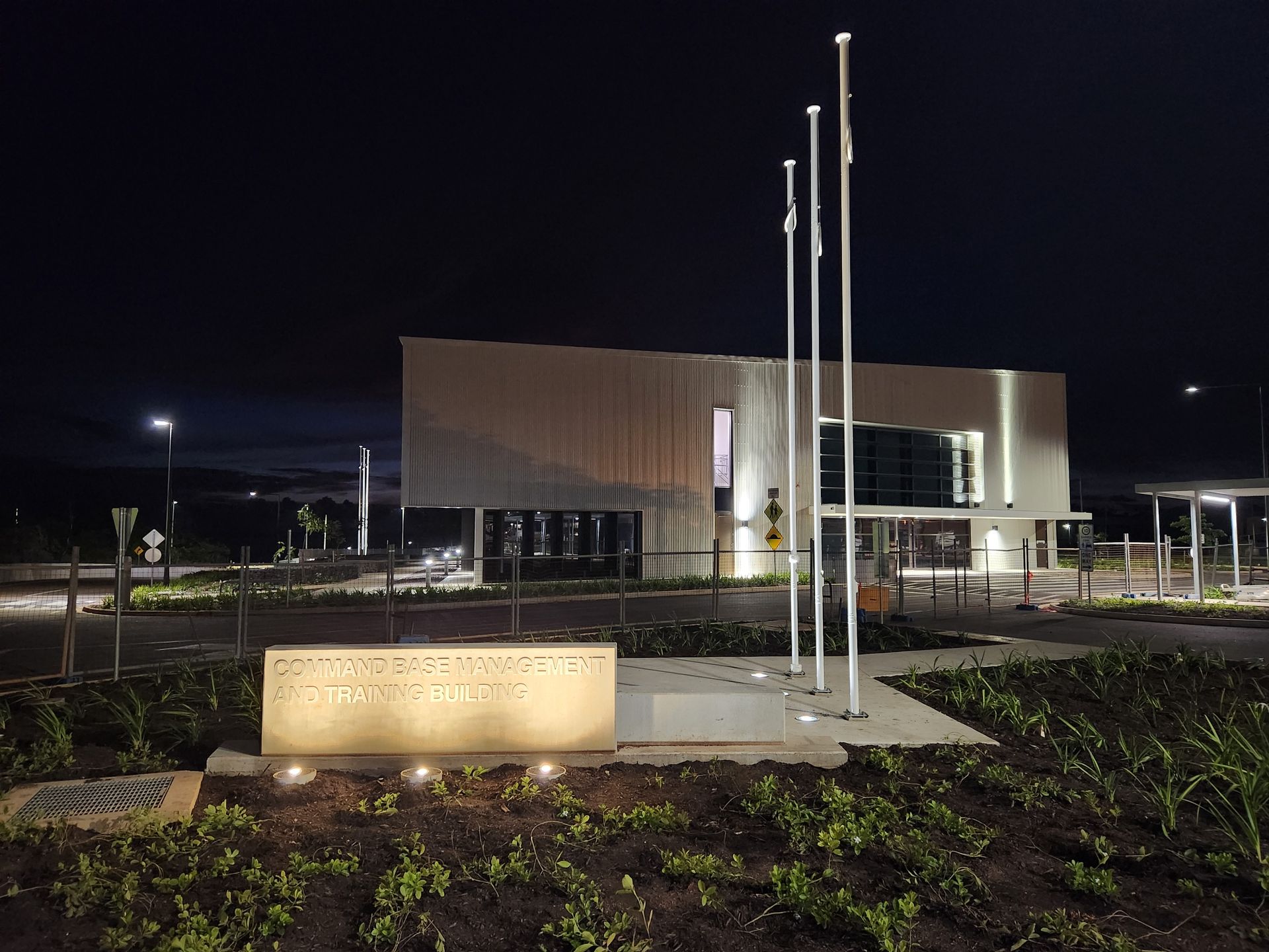 Nighttime view of a modern building with flagpoles, lit windows, and a plaque in front.