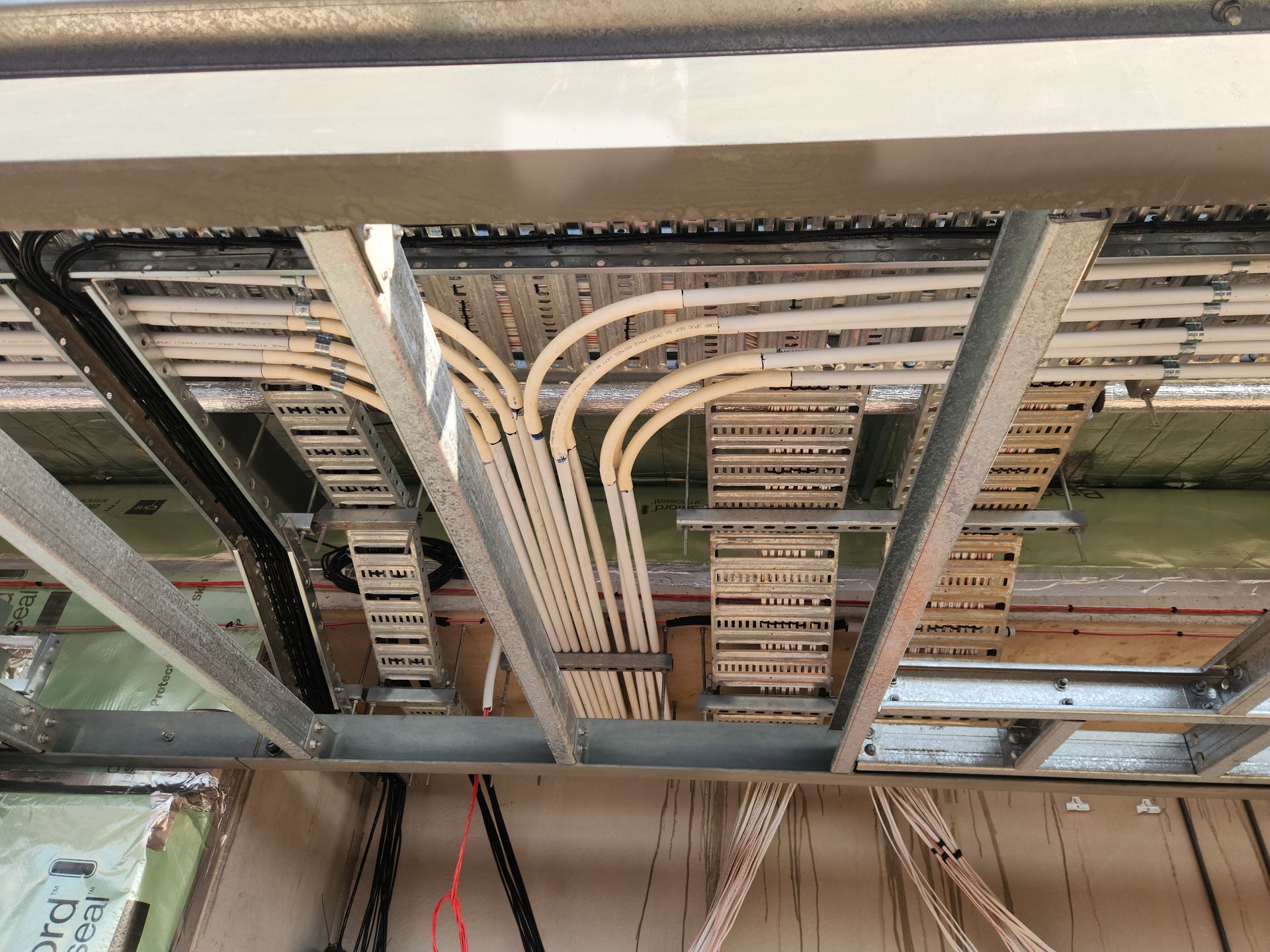 Ceiling of an unfinished building with exposed metal framing, cable trays, and bundled wiring above.