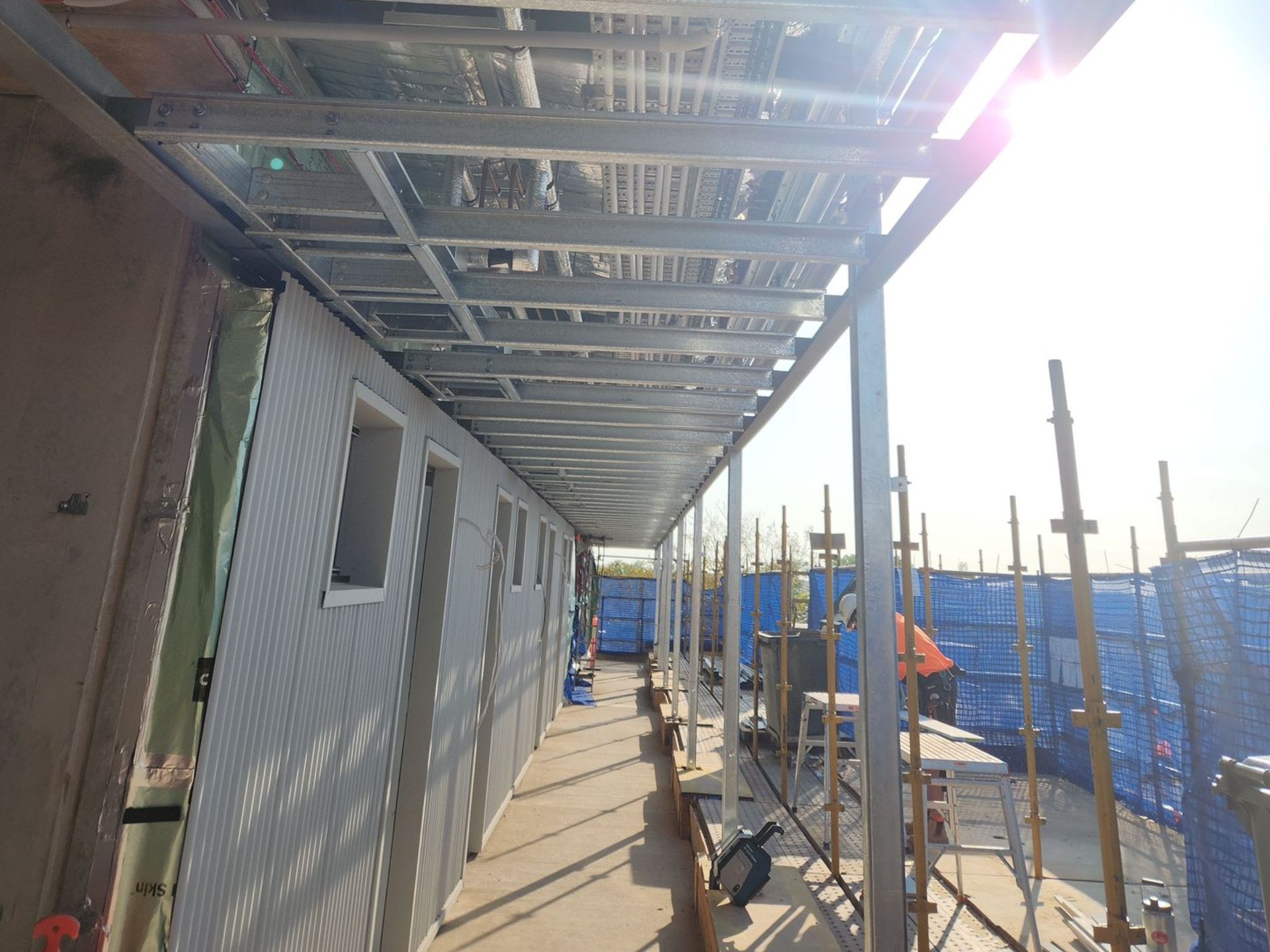 Construction site walkway beside a building, with metal scaffolding overhead and blue safety netting on the right.
