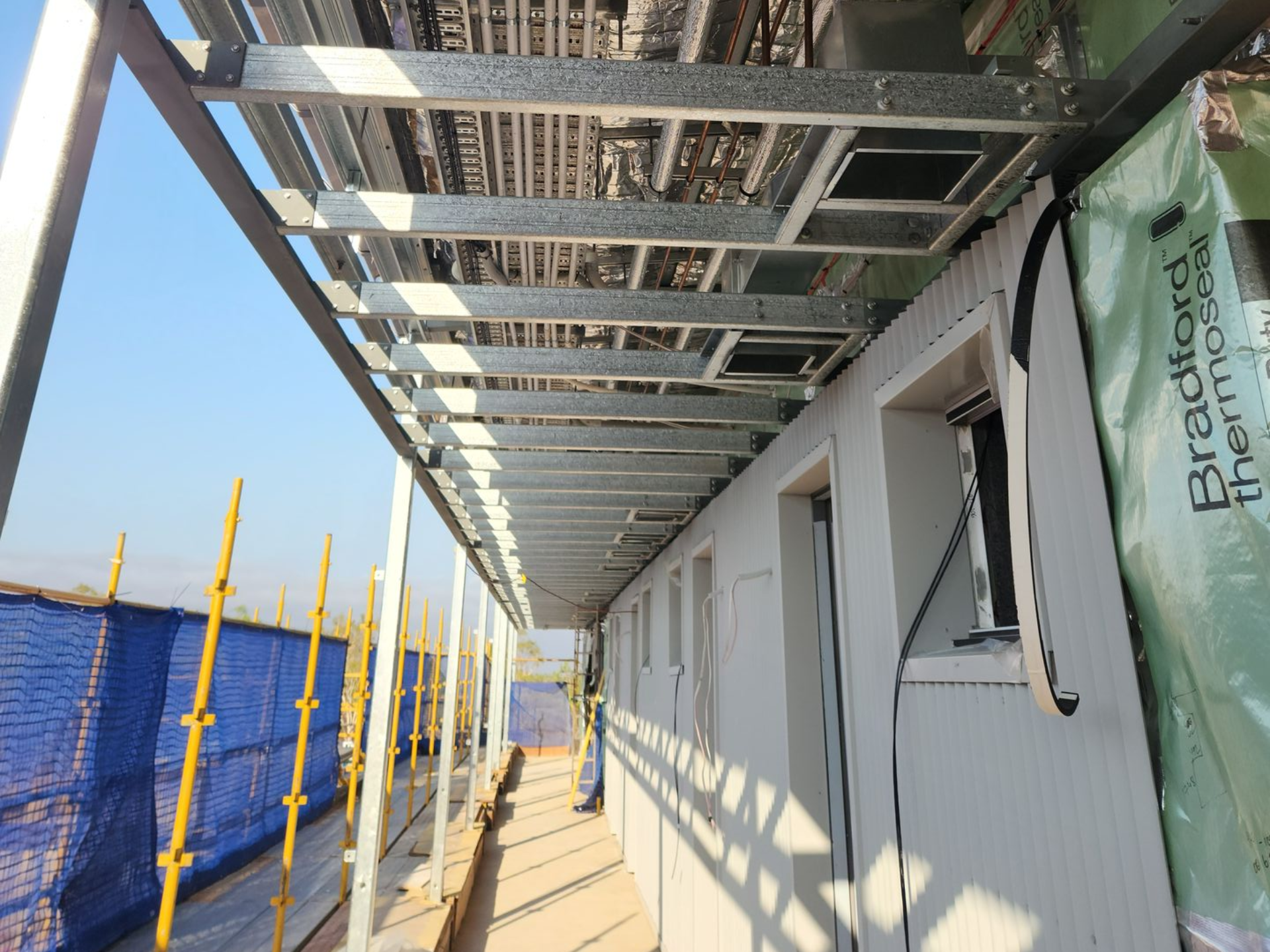 Covered outdoor walkway with metal scaffolding and green house wrap beside a blue construction fence