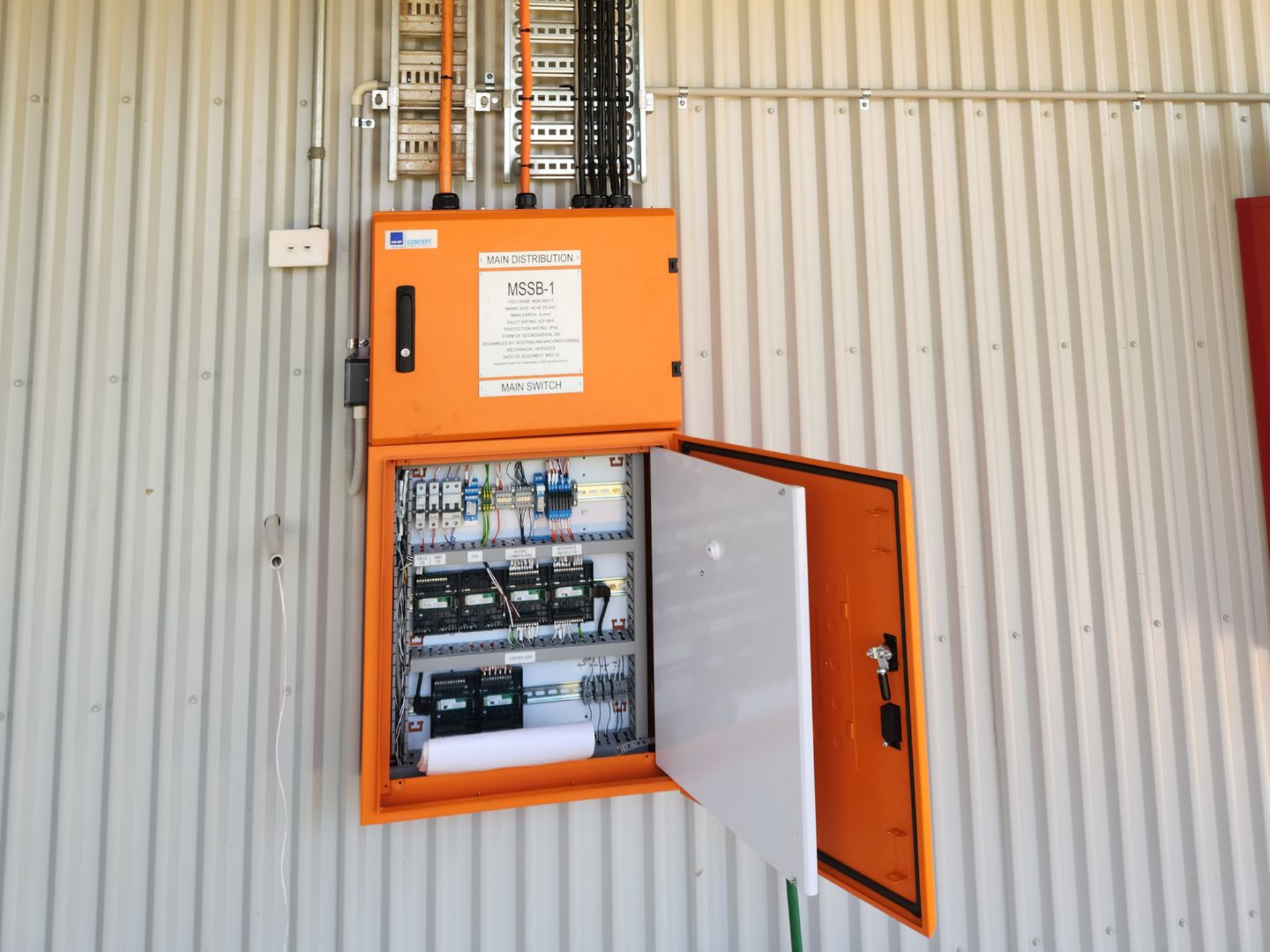 Orange electrical control cabinet open on a corrugated metal wall, showing circuit breakers inside.