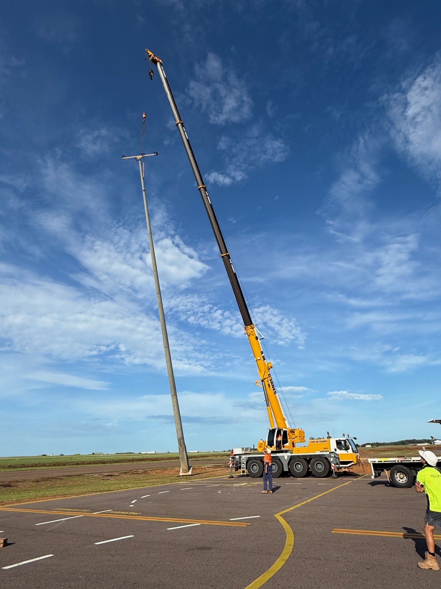 Crane lifting a tall light pole at a road construction site under a blue sky