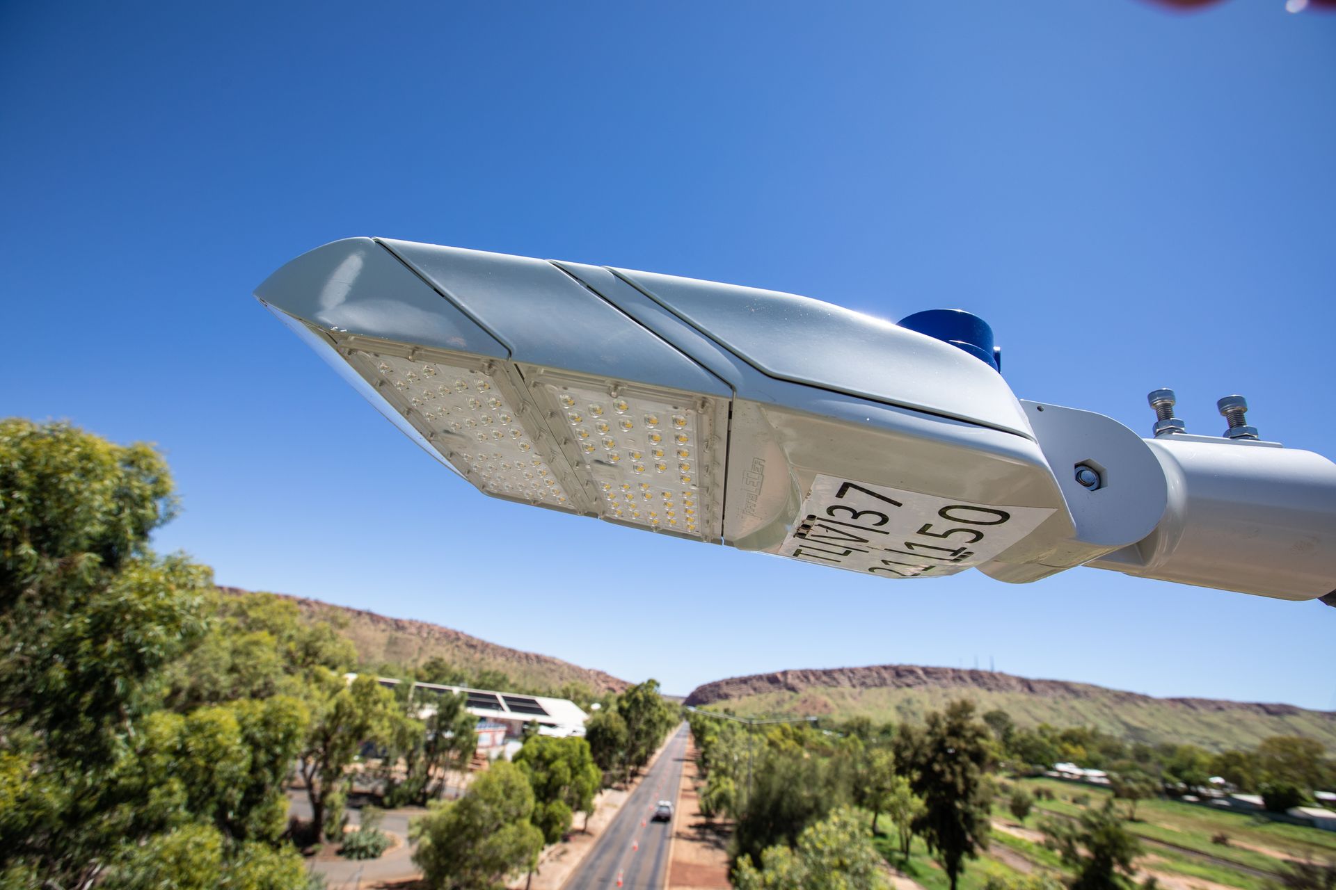 Streetlight close-up over a sunny rural road with trees and hills under a blue sky