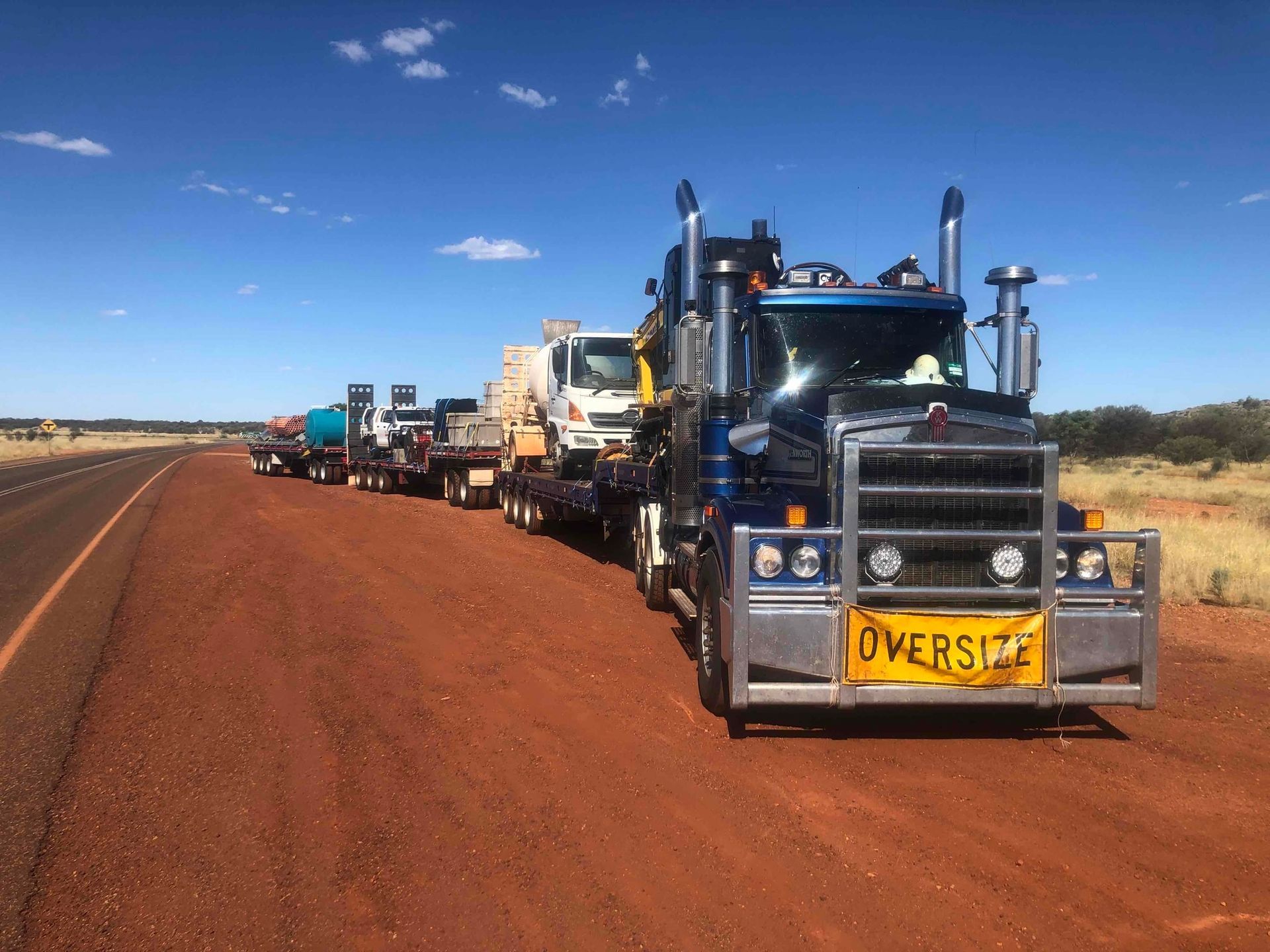 Line of heavy trucks on a red dirt road under a blue sky, lead truck marked “OVERSIZE”