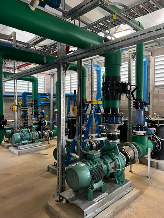 Industrial pump room with green piping and large blue-green pumps on a metal floor.