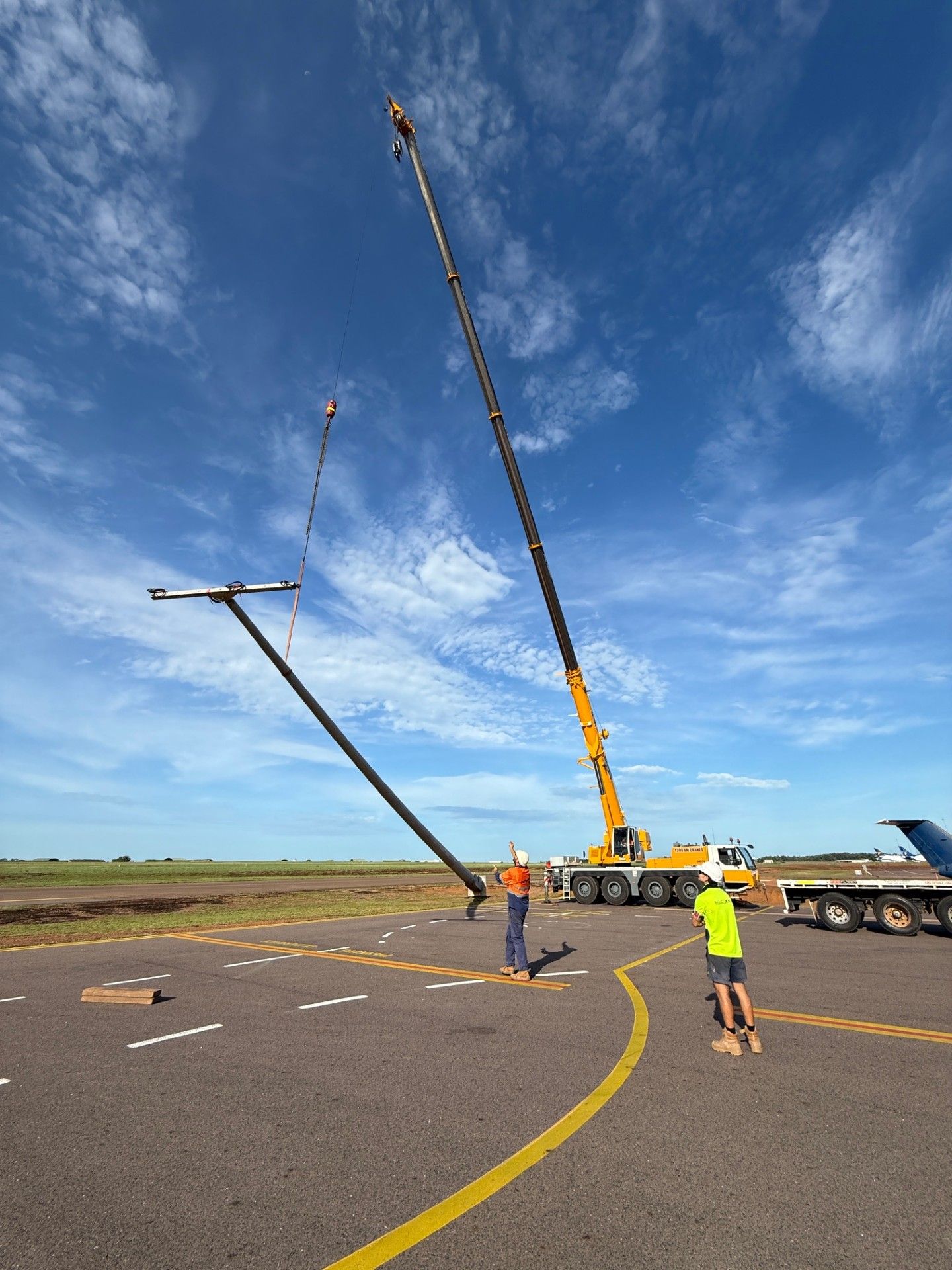 Construction crane lifting a long pole on an airfield, with workers on the tarmac under a blue sky