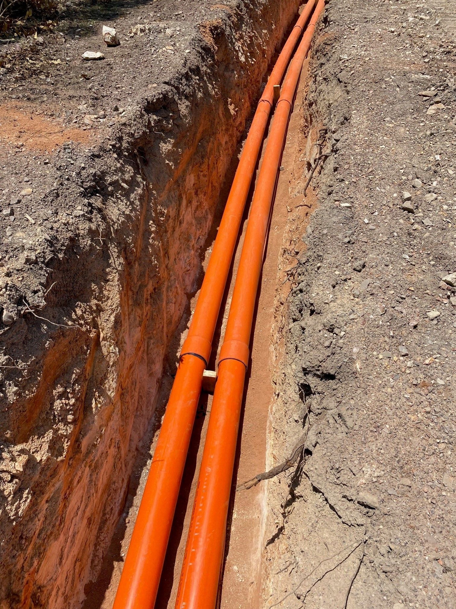 Orange utility pipes laid in a trench beside cracked pavement and exposed dirt.