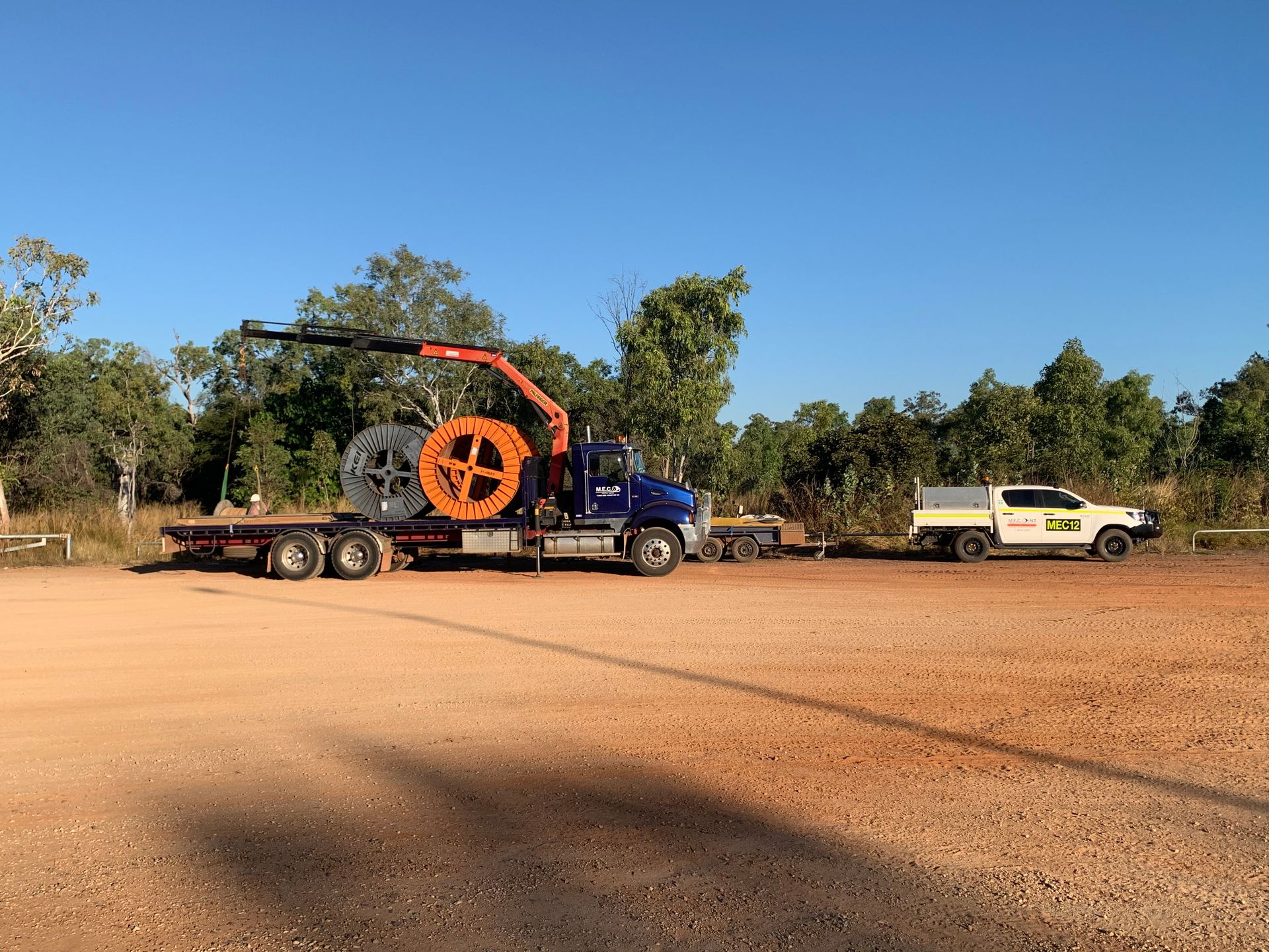 Blue truck with orange crane hauling equipment on a dirt lot, with a white pickup parked beside it.
