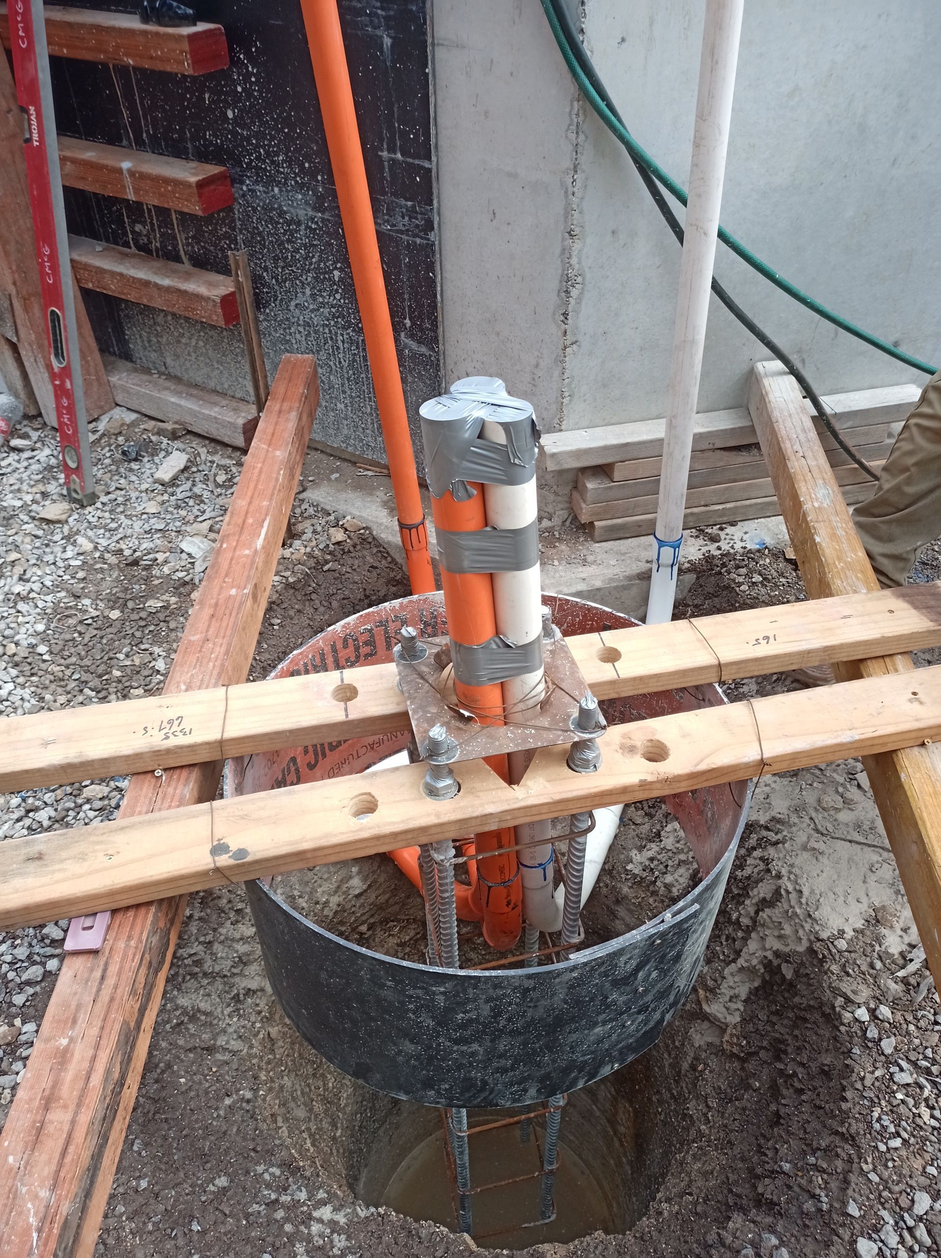 Construction site with a metal cylinder in a hole, supported by wooden planks and orange piping.