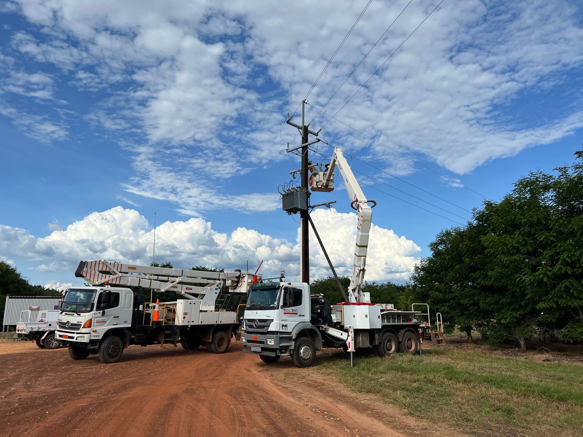 Two utility trucks and a bucket truck working on power lines along a dirt road under a cloudy sky