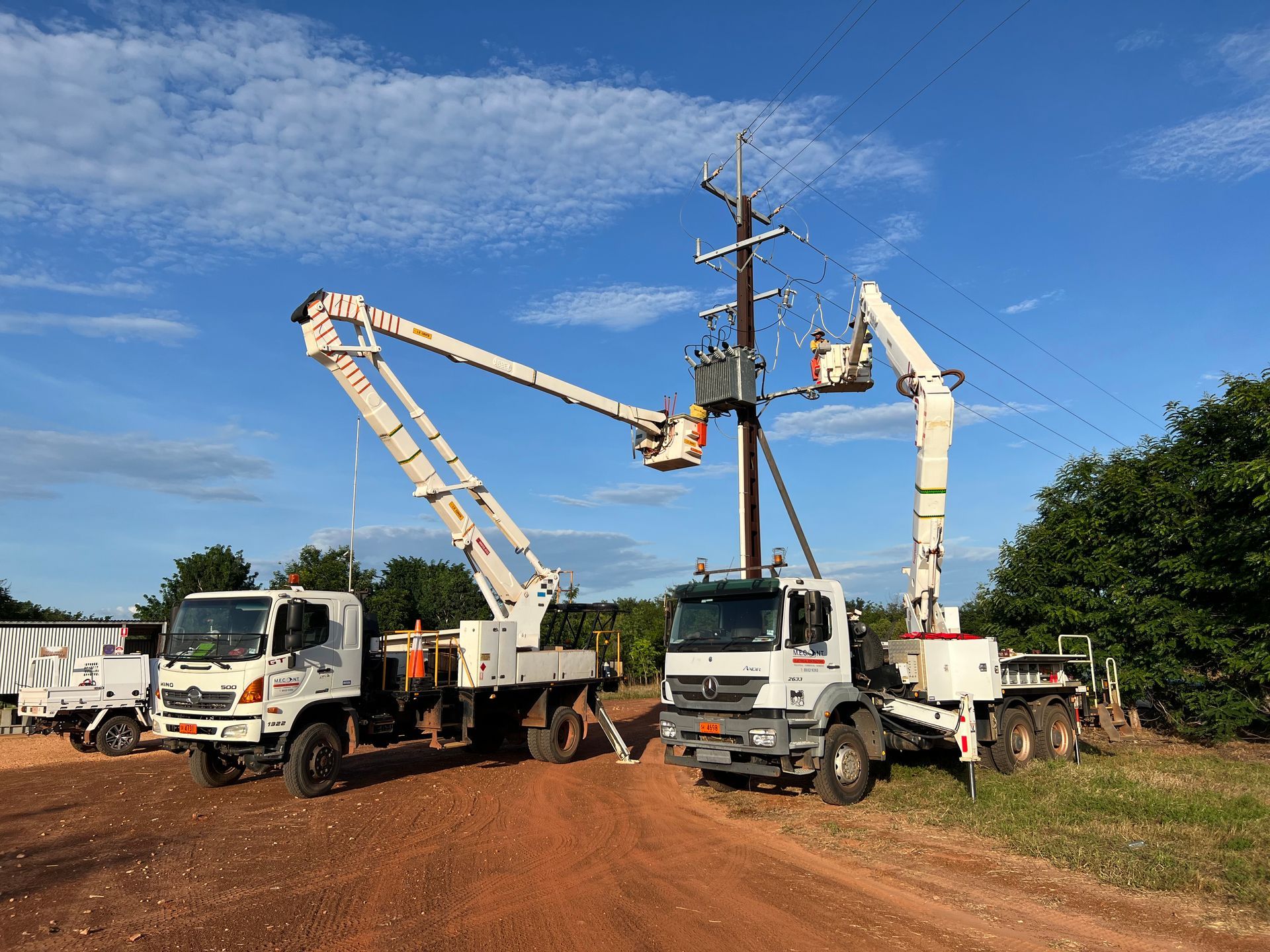 Two bucket trucks and a cherry picker working on power lines beside a dirt road under a blue sky