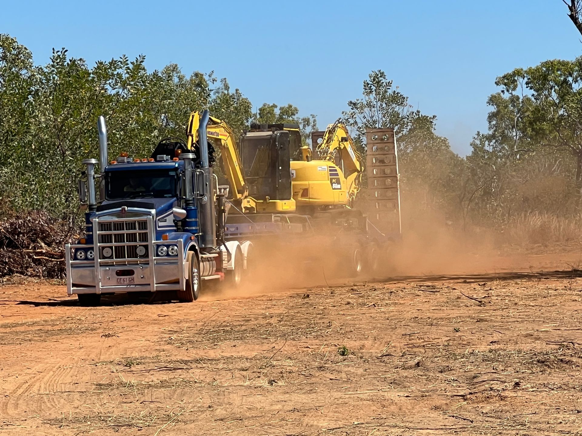 Dump truck hauling excavator along dusty dirt road under clear blue sky