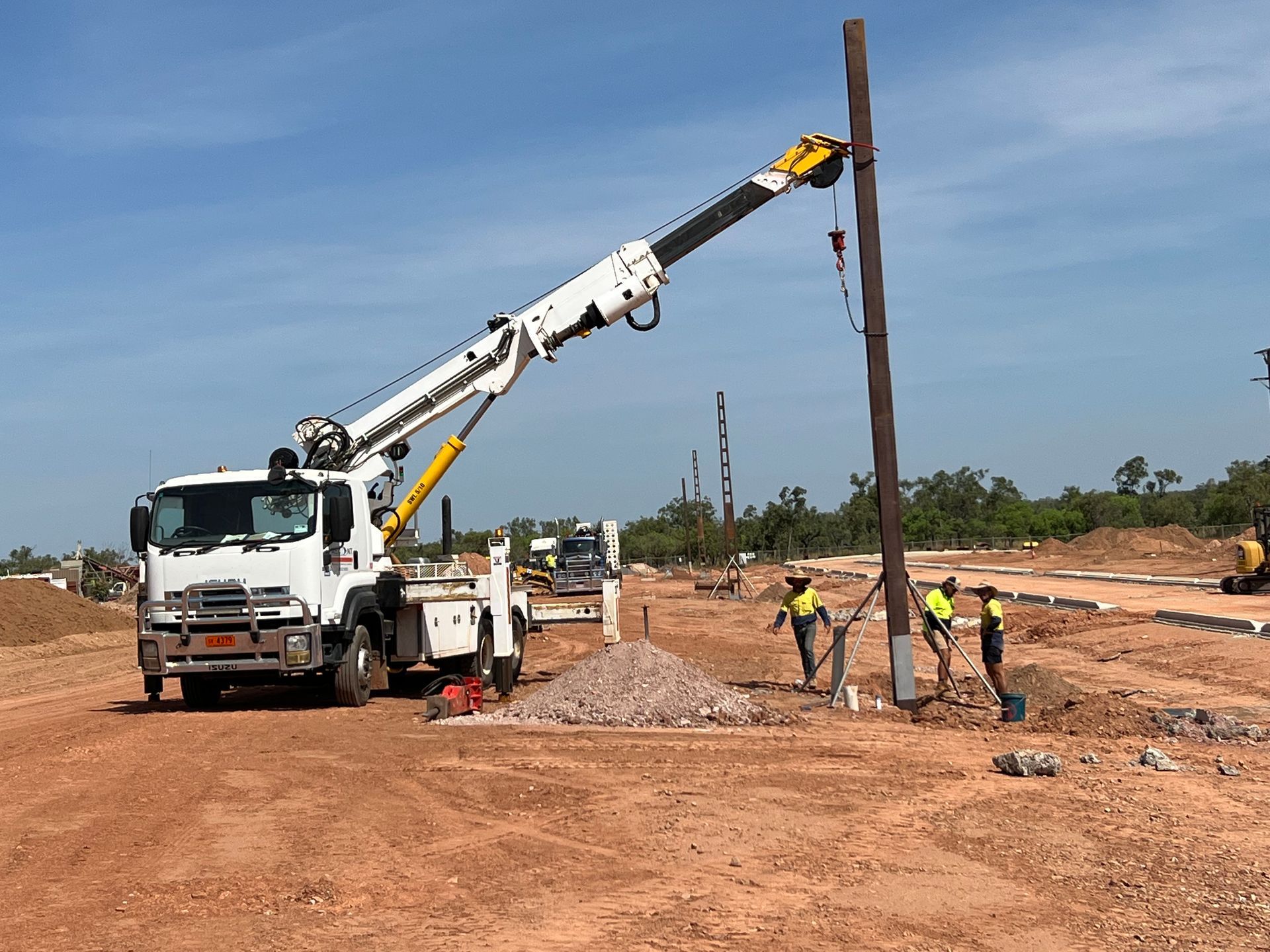 Crane truck lifting a tall utility pole at a road construction site with workers nearby