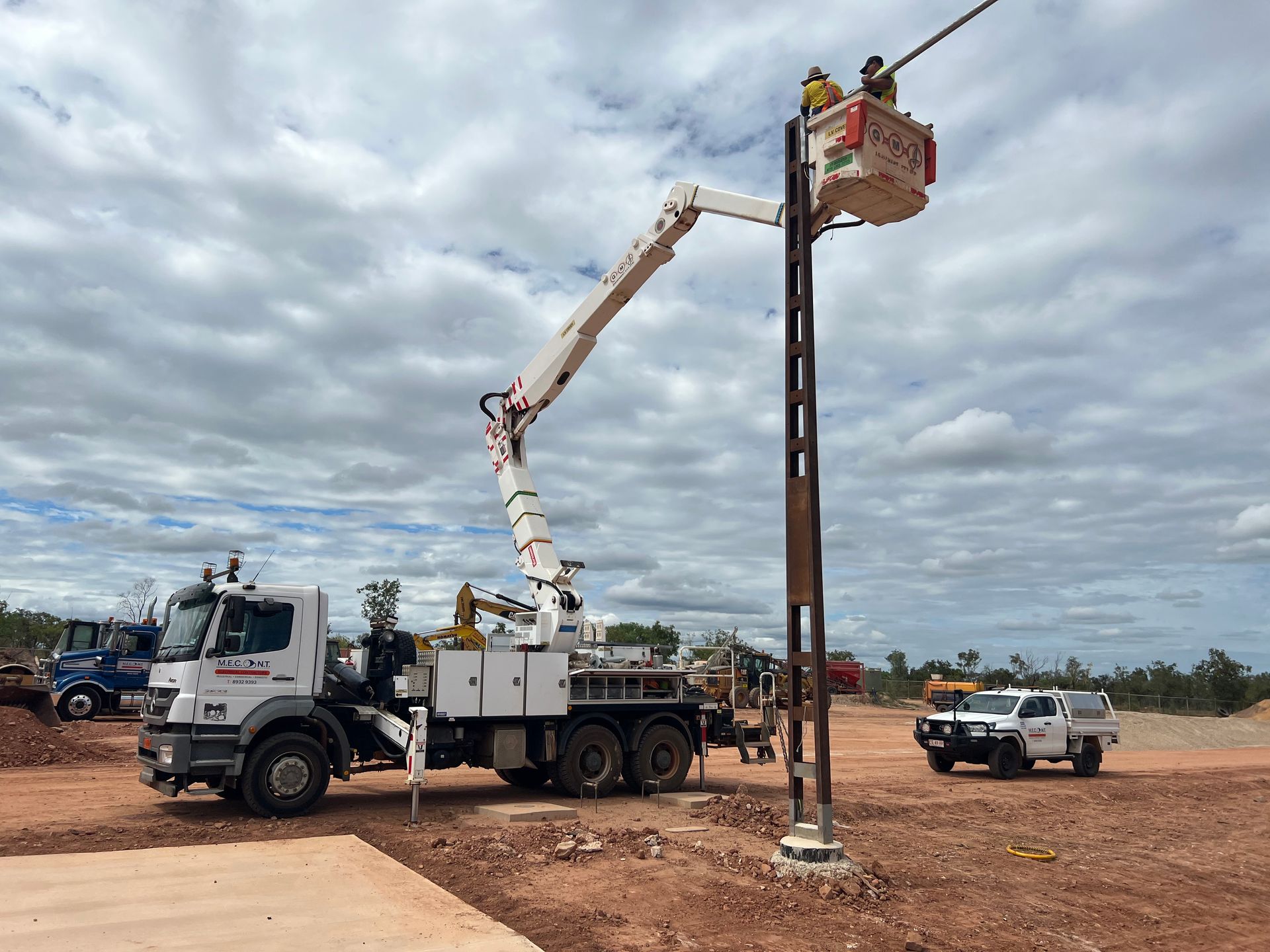 Utility truck with boom lift raising a worker to a pole on a dirt roadside under cloudy skies