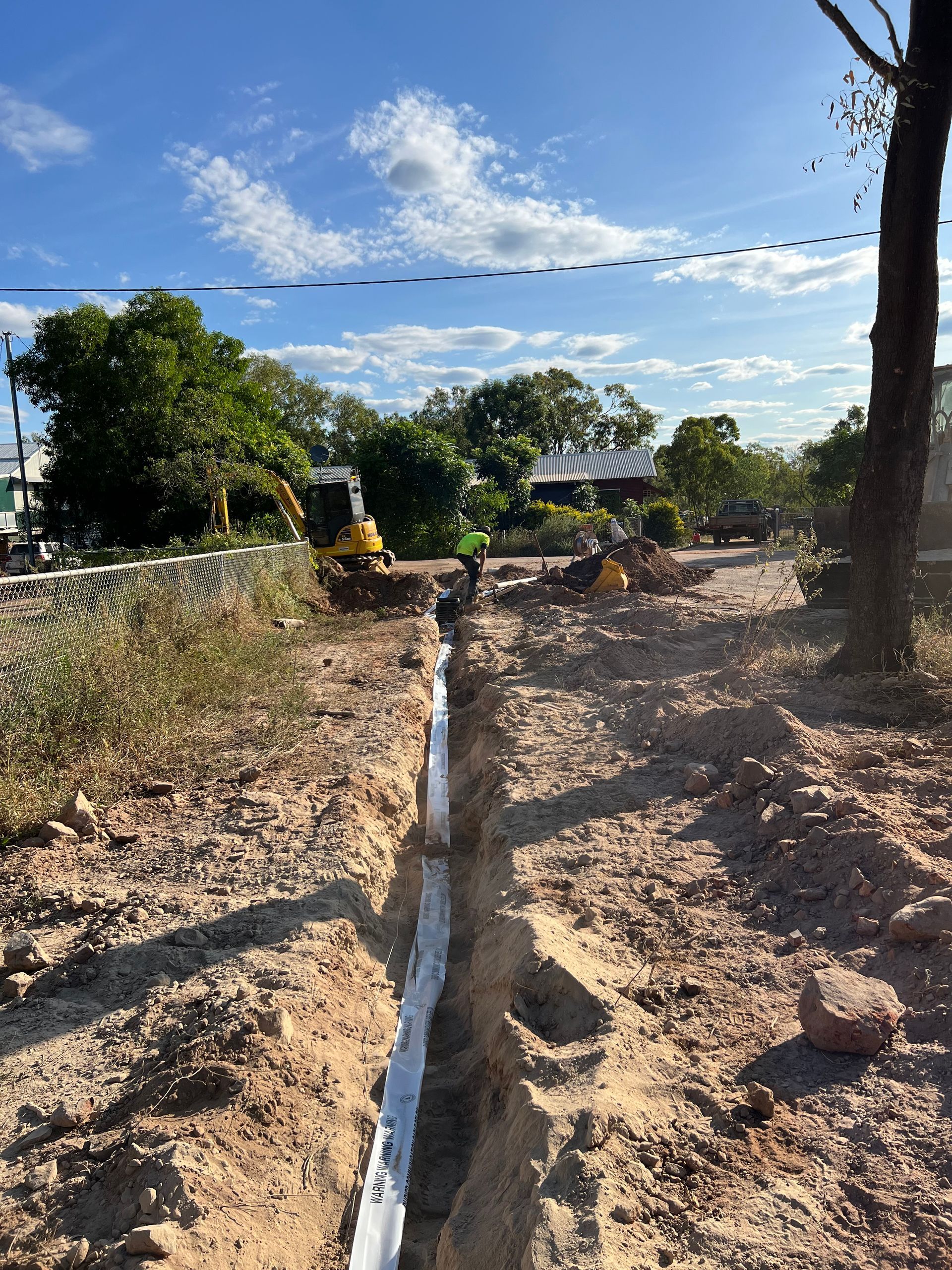 Dirt trench under construction with white lining, trees, and a blue sky with clouds.