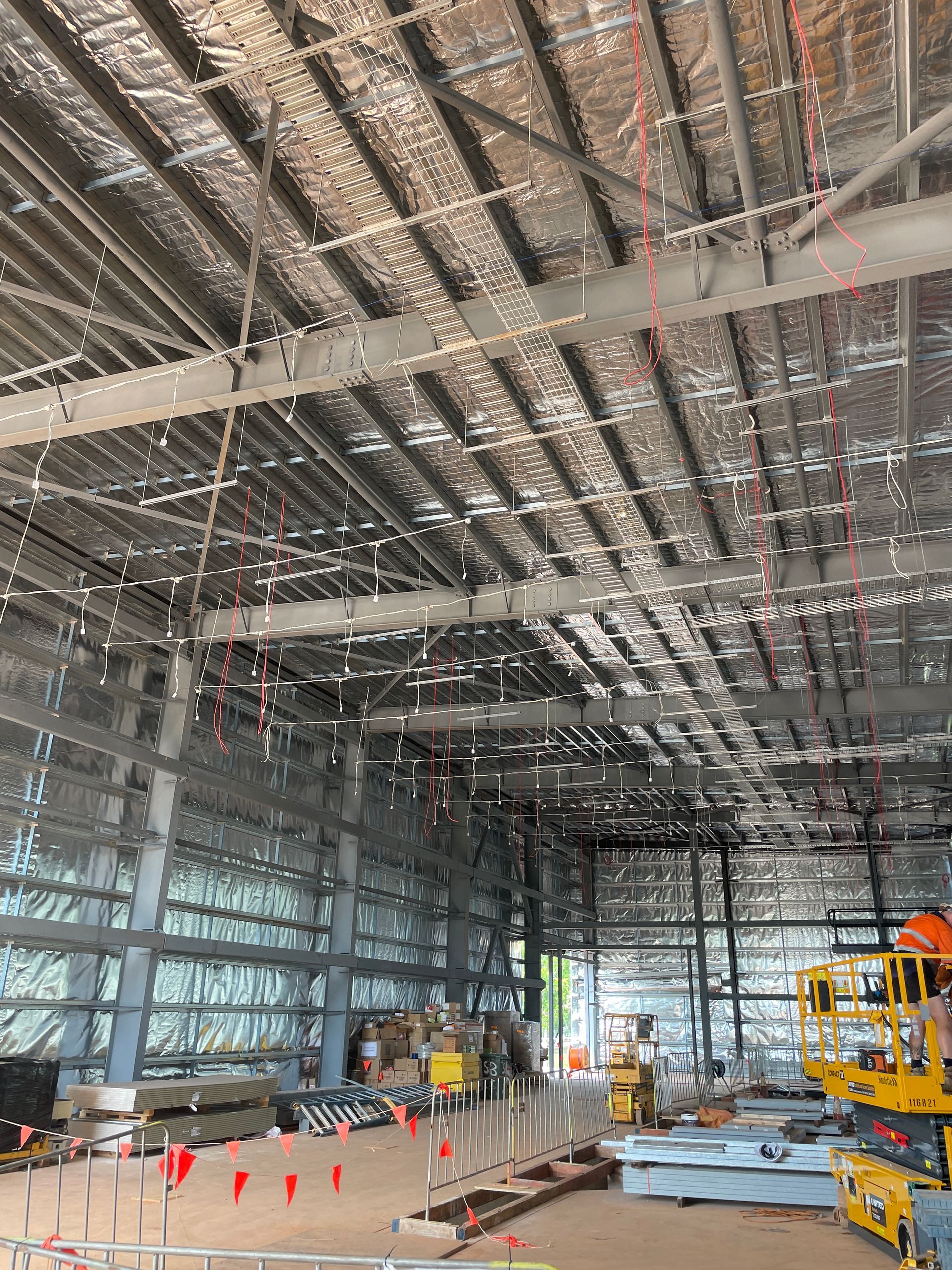Large industrial warehouse interior under construction with exposed ceiling beams, hanging wires, and equipment below