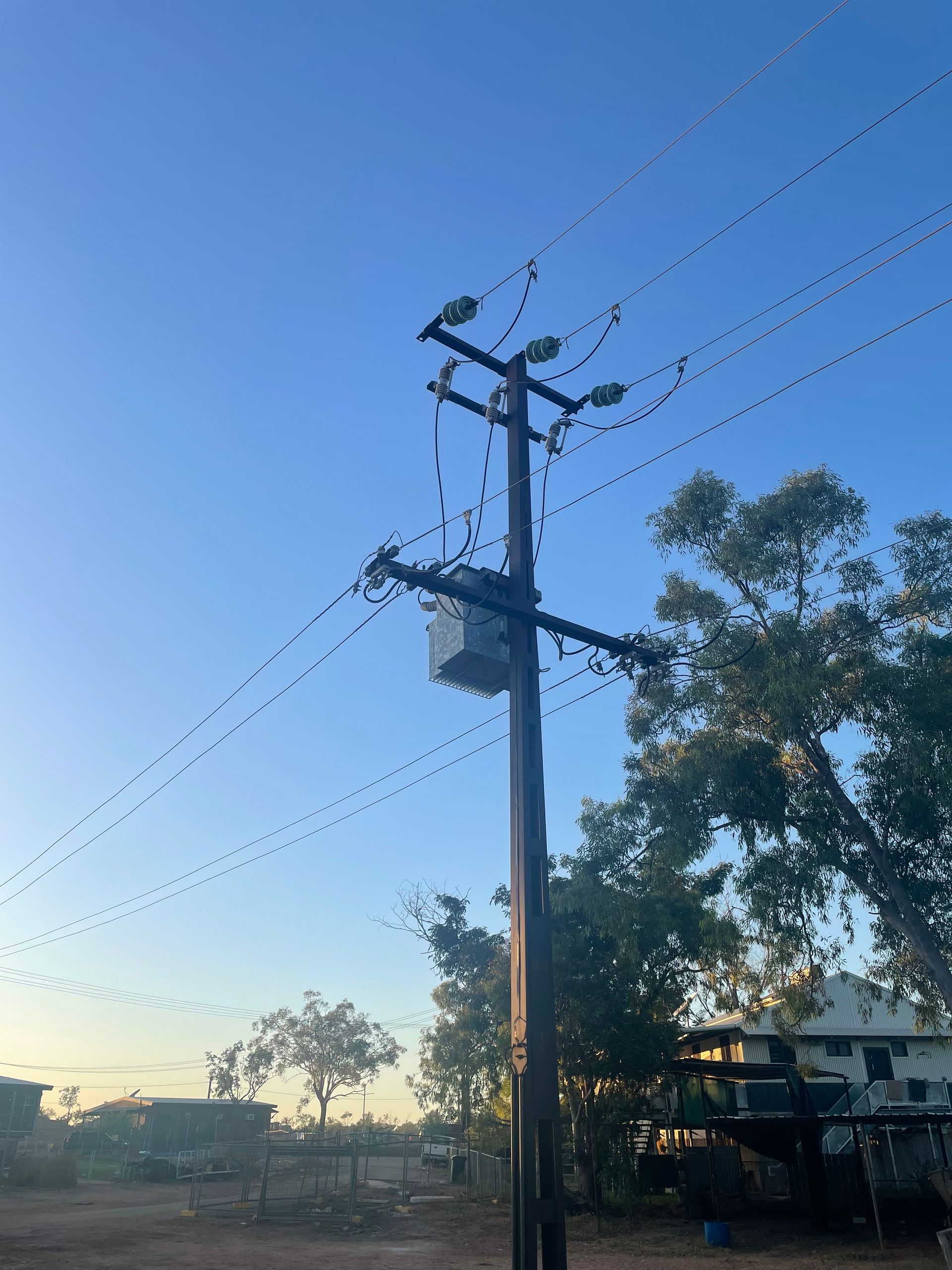 Utility pole with power lines at sunset beside trees and a house