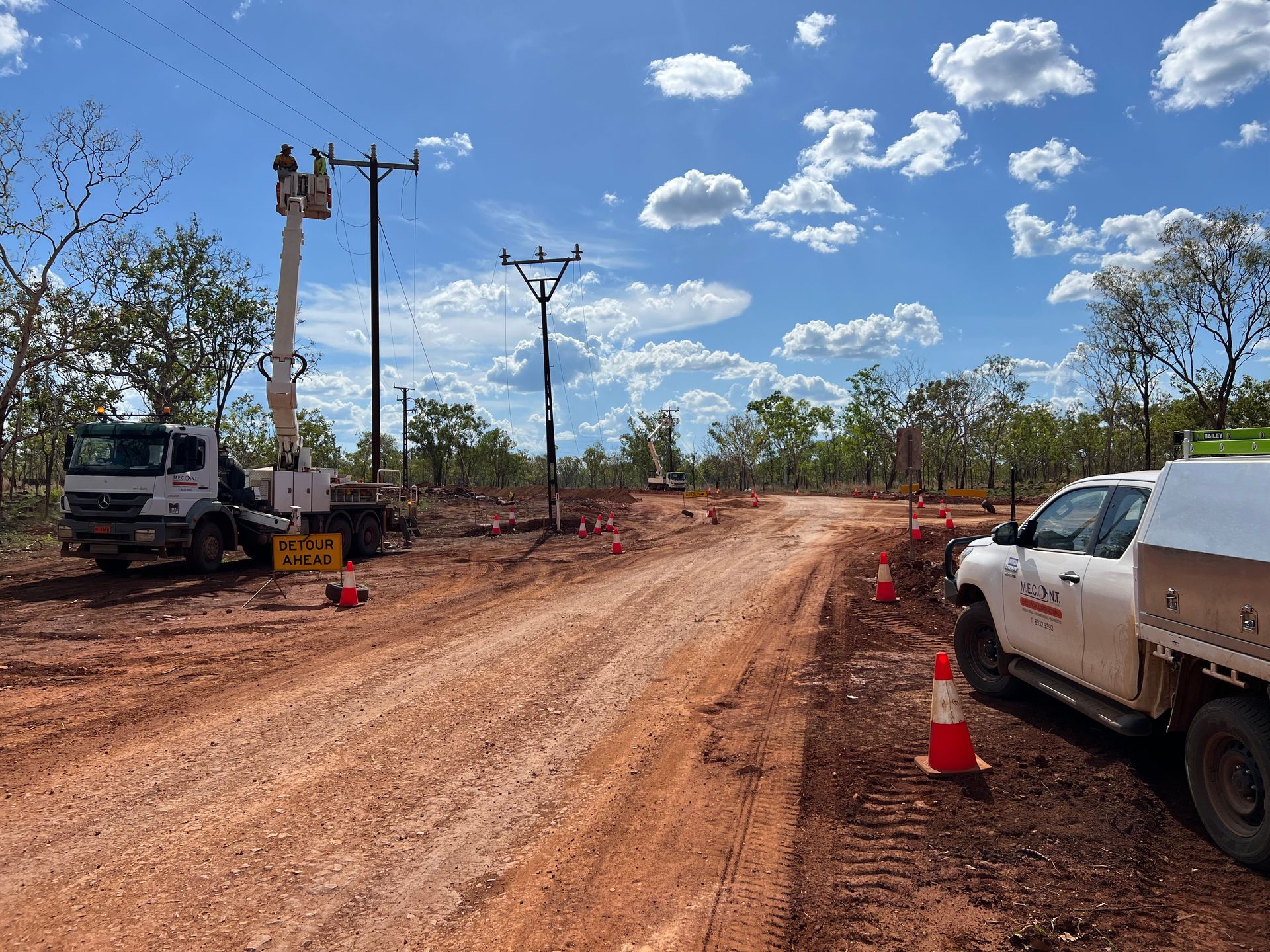 Dirt road construction site with utility trucks, power poles, and orange cones under a blue sky