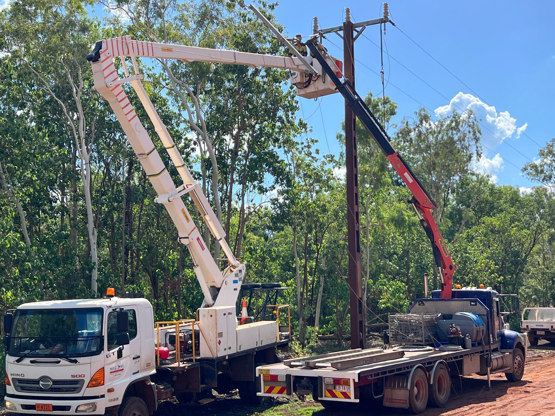 Two utility trucks with raised bucket booms working along a tree-lined roadside under a blue sky.