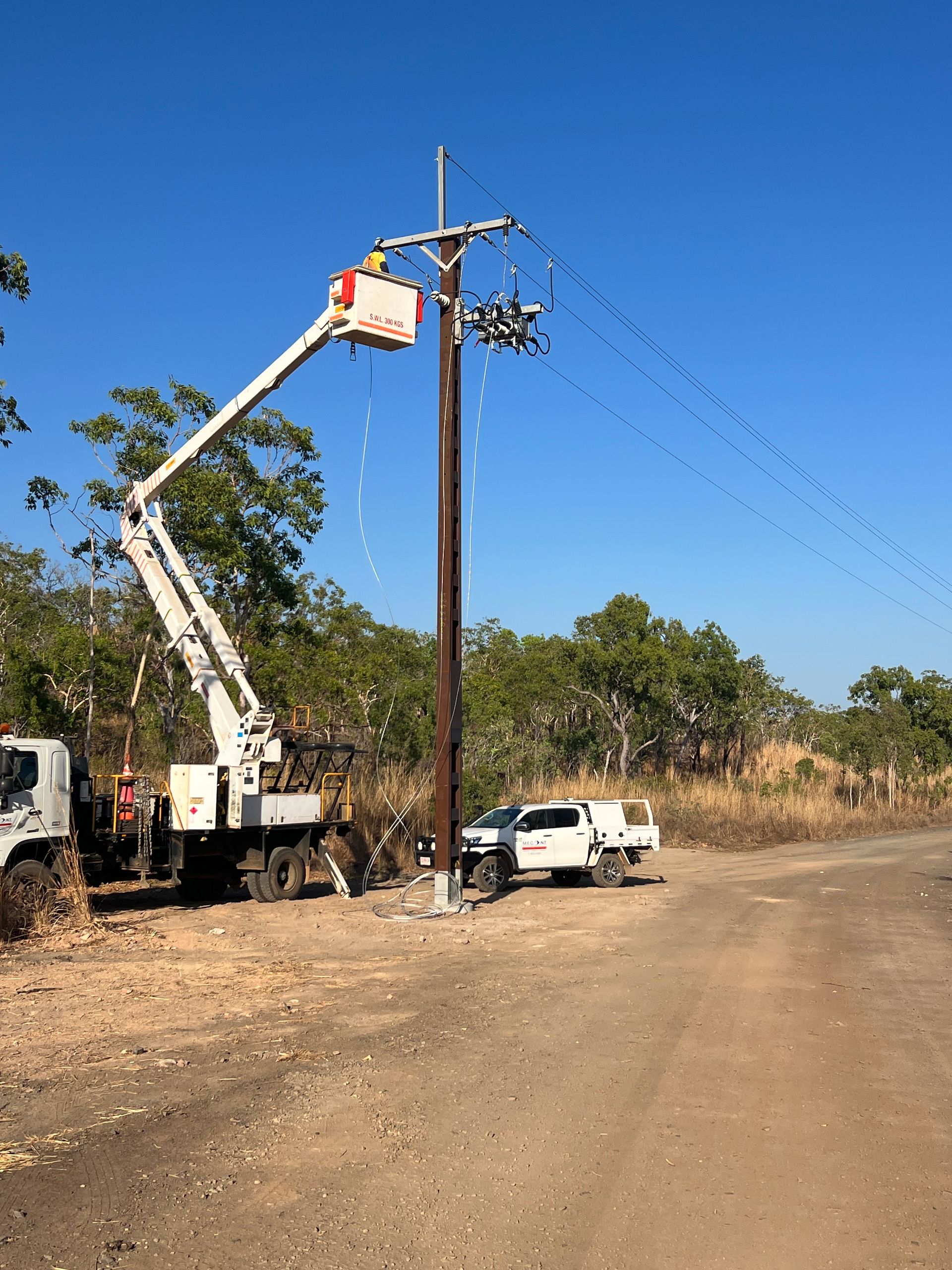 Utility truck with raised bucket working on power lines beside a dirt road under a clear blue sky