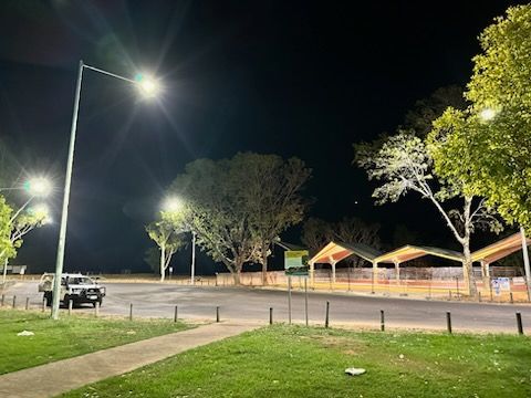 Nighttime park or parking lot with bright streetlights, trees, a small pavilion, and a parked car.