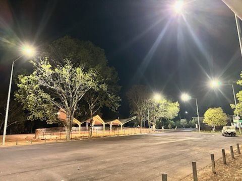 Empty parking lot at night with bright streetlights, trees, and a long fence along the left side.