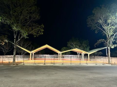 Night view of a lit pavilion with triangular roof frames beside an empty road and trees
