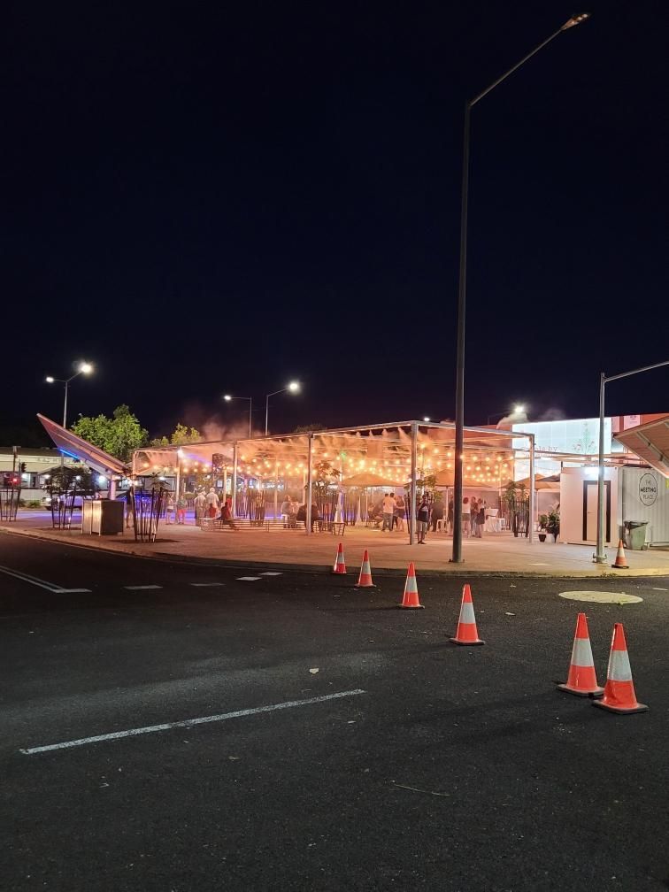 Nighttime street scene with a lit outdoor dining area, traffic cones, and a parked car by the road.